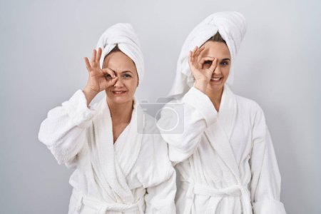 Téléchargez les photos : Femme et fille d'âge moyen portant un peignoir blanc et une serviette faisant un geste correct avec le sourire de la main, les yeux regardant à travers les doigts avec un visage heureux. - en image libre de droit