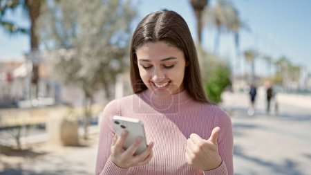 Téléchargez les photos : Jeune belle femme hispanique à l'aide d'un smartphone faisant un geste pouce vers le haut au parc - en image libre de droit