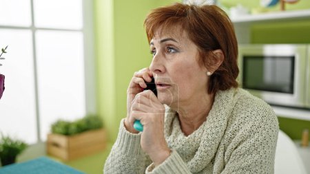 Téléchargez les photos : Mature hispanique femme tenant respirateur vape assis sur la table parlant au téléphone à la salle à manger - en image libre de droit