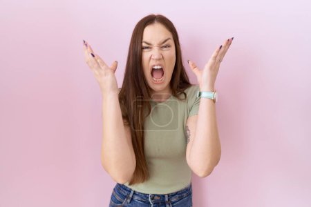 Photo for Beautiful brunette woman standing over pink background crazy and mad shouting and yelling with aggressive expression and arms raised. frustration concept. - Royalty Free Image