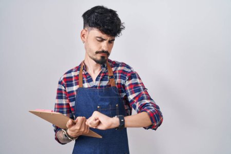 Téléchargez les photos : Jeune homme hispanique avec barbe portant tablier de serveur tenant presse-papiers vérifier l'heure sur la montre-bracelet, détendu et confiant - en image libre de droit