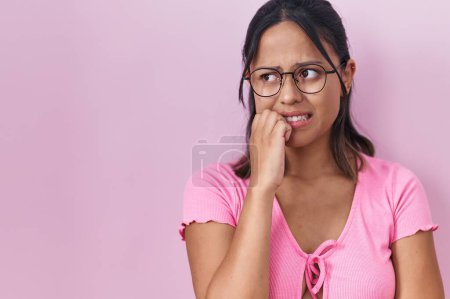 Foto de Mujer joven hispana de pie sobre fondo rosa con gafas que se ven estresadas y nerviosas con las manos en la boca mordiendo uñas. problema de ansiedad. - Imagen libre de derechos