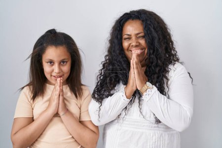 Foto de Madre e hija joven de pie sobre fondo blanco rezando con las manos juntas pidiendo perdón sonriendo confiadas. - Imagen libre de derechos