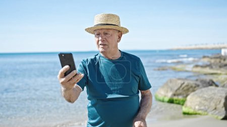 Téléchargez les photos : Homme âgé aux cheveux gris touriste portant un chapeau d'été en utilisant un smartphone au bord de la mer - en image libre de droit