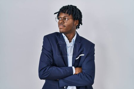 Téléchargez les photos : Jeune homme africain avec dreadlocks portant une veste d'affaires sur fond blanc regardant du côté avec les bras croisés convaincu et confiant - en image libre de droit