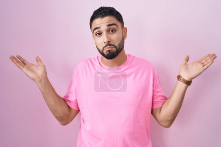 Photo for Hispanic young man standing over pink background clueless and confused expression with arms and hands raised. doubt concept. - Royalty Free Image