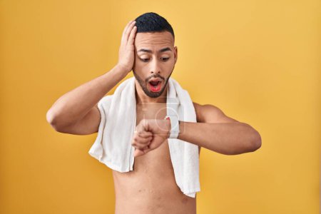 Foto de Joven hombre hispano de pie sin camisa con toalla mirando el tiempo del reloj preocupado, con miedo de llegar tarde - Imagen libre de derechos