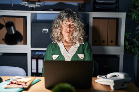 Téléchargez les photos : Femme d'âge moyen travaillant la nuit à l'aide d'un ordinateur portable détendu avec une expression sérieuse sur le visage. simple et naturel regardant la caméra. - en image libre de droit