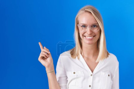Téléchargez les photos : Jeune femme caucasienne debout sur fond bleu avec un grand sourire sur le visage, pointant avec la main et le doigt vers le côté regardant la caméra. - en image libre de droit