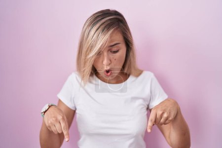 Photo for Young blonde woman standing over pink background pointing down with fingers showing advertisement, surprised face and open mouth - Royalty Free Image