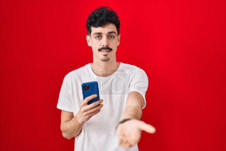 Foto de Hombre hispano usando smartphone sobre fondo rojo sonriente alegre ofreciendo mano de palma dando asistencia y aceptación. - Imagen libre de derechos