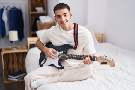 Téléchargez les photos : Jeune homme hispanique jouant de la guitare électrique assis sur le lit dans la chambre - en image libre de droit