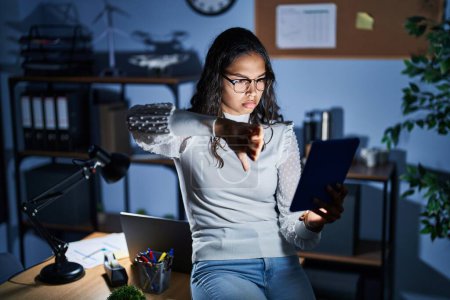 Photo for Young brazilian woman using touchpad at night working at the office looking unhappy and angry showing rejection and negative with thumbs down gesture. bad expression. - Royalty Free Image