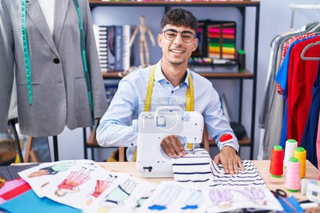 Foto de Joven hombre hispano sastre sonriendo confiado usando máquina de coser en sastrería - Imagen libre de derechos