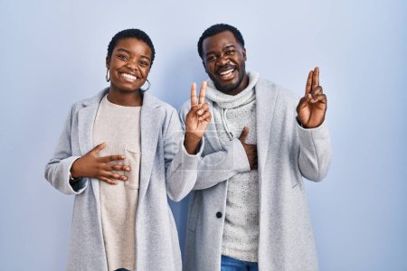 Foto de Joven pareja afroamericana de pie sobre fondo azul juntos sonriendo jurando con la mano en el pecho y los dedos hacia arriba, haciendo una promesa de lealtad juramento - Imagen libre de derechos