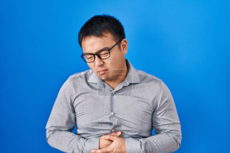 Téléchargez les photos : Jeune homme chinois debout sur fond bleu avec la main sur le ventre parce que les nausées, la maladie douloureuse se sentent mal. concept de douleur. - en image libre de droit