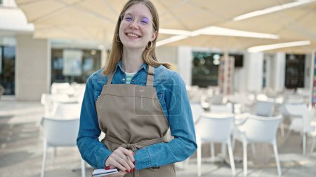 Foto de Joven camarera rubia sonriendo confiada sosteniendo el cuaderno en la terraza de la cafetería - Imagen libre de derechos