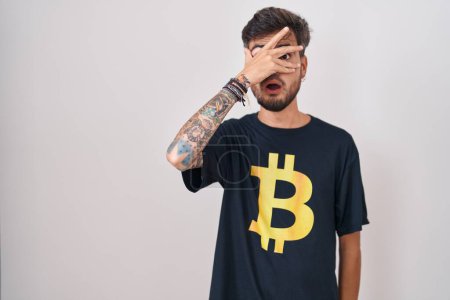 Foto de Joven hombre hispano con tatuajes con camiseta bitcoin asomándose con la mano a la cara y los ojos, mirando a través de los dedos con expresión avergonzada. - Imagen libre de derechos