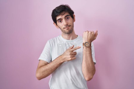 Photo for Young hispanic man standing over pink background in hurry pointing to watch time, impatience, looking at the camera with relaxed expression - Royalty Free Image