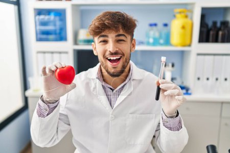 Photo for Arab man with beard working at scientist laboratory holding blood samples smiling and laughing hard out loud because funny crazy joke. - Royalty Free Image