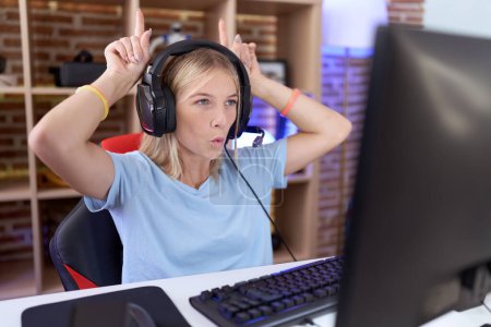 Téléchargez les photos : Jeune femme caucasienne jouant à des jeux vidéo portant un casque faisant un geste drôle avec le doigt sur la tête comme des cornes de taureau - en image libre de droit
