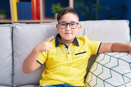 Foto de Joven niño hispano sentado en el sofá en casa sonriendo feliz señalando con la mano y el dedo - Imagen libre de derechos