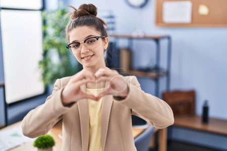 Téléchargez les photos : Adolescente travaillant au bureau portant des lunettes souriant en amour montrant le symbole du cœur et la forme avec les mains. concept romantique. - en image libre de droit