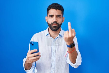Foto de Hispanic man with beard using smartphone typing message showing middle finger, impolite and rude fuck off expression - Imagen libre de derechos