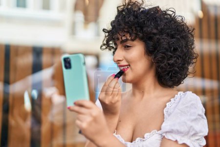Téléchargez les photos : Jeune Moyen-Orient femme maquillage lèvres regarder smartphone à la rue - en image libre de droit