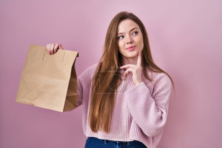 Photo for Young caucasian woman holding shopping bag and credit card serious face thinking about question with hand on chin, thoughtful about confusing idea - Royalty Free Image