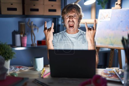 Photo for Middle age man sitting at art studio with laptop at night shouting with crazy expression doing rock symbol with hands up. music star. heavy concept. - Royalty Free Image