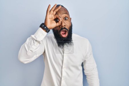 Photo for African american man standing over blue background doing ok gesture shocked with surprised face, eye looking through fingers. unbelieving expression. - Royalty Free Image