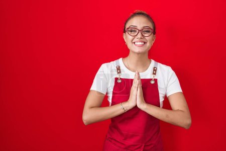 Photo for Young hispanic woman wearing waitress apron over red background praying with hands together asking for forgiveness smiling confident. - Royalty Free Image