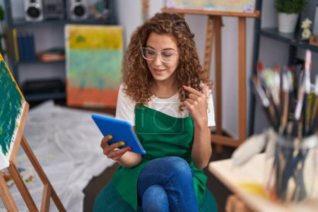Foto de Joven mujer caucásica pintor sentado en el estudio de arte haciendo videollamada con la tableta sonriendo feliz señalando con la mano y el dedo - Imagen libre de derechos