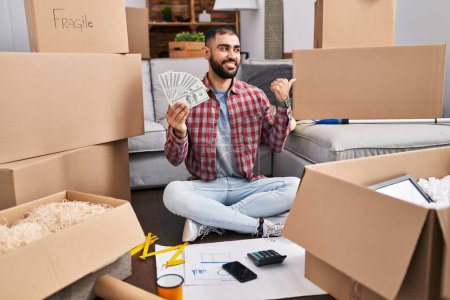 Photo for Middle east man with beard sitting on the floor at new home holding money pointing thumb up to the side smiling happy with open mouth - Royalty Free Image