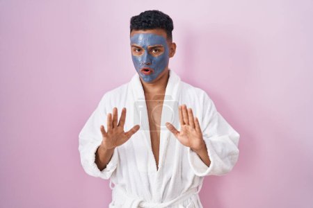 Photo for Young hispanic man wearing beauty face mask and bath robe moving away hands palms showing refusal and denial with afraid and disgusting expression. stop and forbidden. - Royalty Free Image