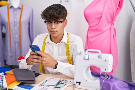 Téléchargez les photos : Jeune adolescent hispanique tailleur à l'aide d'un smartphone au magasin de tailleur - en image libre de droit