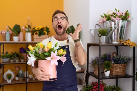 Foto de Hombre de mediana edad con barba trabajando en floristería sosteniendo planta celebrando la victoria con sonrisa feliz y expresión ganadora con las manos levantadas - Imagen libre de derechos