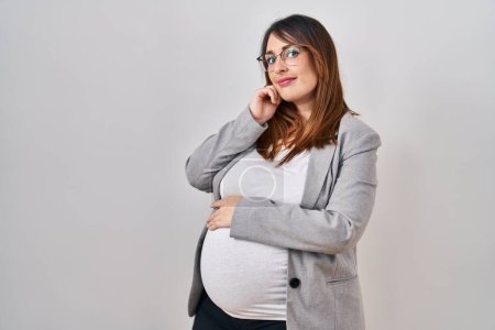Photo for Pregnant business woman standing over white background serious face thinking about question with hand on chin, thoughtful about confusing idea - Royalty Free Image