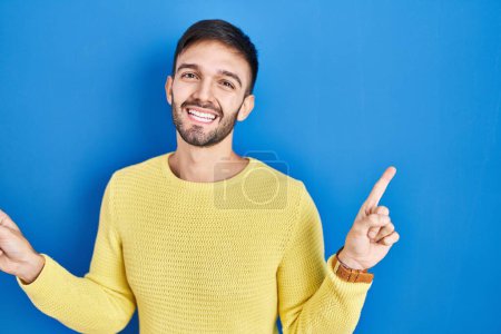 Téléchargez les photos : Homme hispanique debout sur fond bleu souriant confiant pointant avec les doigts vers différentes directions. espace de copie pour la publicité - en image libre de droit