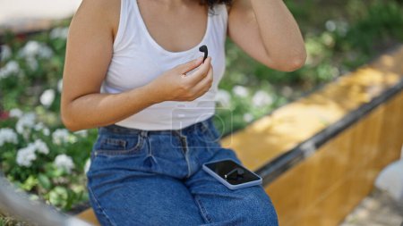 Téléchargez les photos : Jeune belle femme hispanique assise sur un banc tenant écouteurs et smartphone au parc - en image libre de droit
