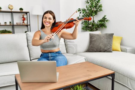 Photo for Young beautiful hispanic woman having online violin class sitting on sofa at home - Royalty Free Image