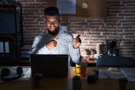 Photo for Young hispanic man working at the office at night smiling and looking at the camera pointing with two hands and fingers to the side. - Royalty Free Image
