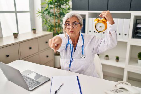 Foto de Mujer de mediana edad con cabello gris vistiendo uniforme médico sosteniendo el despertador apuntando con el dedo a la cámara y a ti, gesto confiado que se ve serio - Imagen libre de derechos