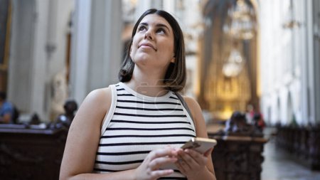 Téléchargez les photos : Jeune belle femme hispanique visitant l'église en utilisant un smartphone debout à l'église augustinienne de Vienne - en image libre de droit