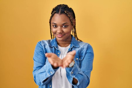Téléchargez les photos : Femme afro-américaine avec des tresses debout sur fond jaune souriant avec les mains paumes ensemble recevant ou donnant geste. maintien et protection - en image libre de droit