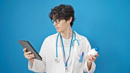 Foto de Joven médico hispano usando touchpad sosteniendo frasco de medicina sobre fondo azul aislado - Imagen libre de derechos