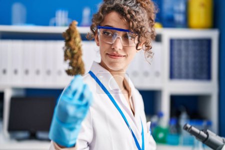 Téléchargez les photos : Young hispanic woman scientist smiling confident holding marihuana plant at laboratory - en image libre de droit