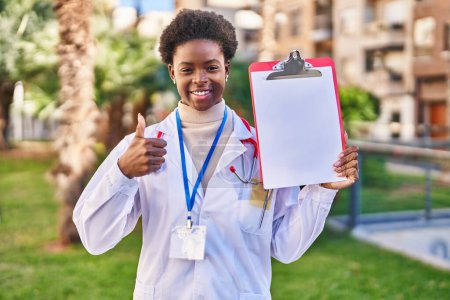 Photo for African american woman wearing doctor stethoscope holding clipboard smiling happy and positive, thumb up doing excellent and approval sign - Royalty Free Image