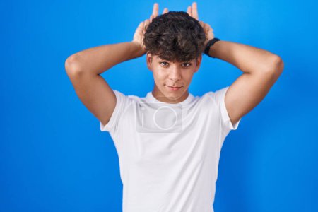 Téléchargez les photos : Adolescent hispanique debout sur fond bleu posant drôle et fou avec les doigts sur la tête comme des oreilles de lapin, souriant joyeux - en image libre de droit